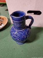 Blue pottery vase with carved raised relief work, handle and small spout, shown from side angle