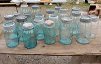 Photo of 15 glass Mason jars with blue tint arranged on a wooden surface outdoors. Various sizes and lid conditions, metal lids present on all jars.