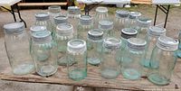 Twenty glass mason jars with metal lids, varying in size and showing green tint, arranged on wooden surface for display