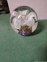 Clear round glass paperweight with internal white floral design and colorful base on green surface