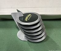 Stacked view of six round metal Jameson's whiskey coasters in curved metal holder, showing brushed metal edges and black center with yellow Jameson's logo