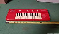 Front view of the bright red Radio Shack electric organ showing keyboard, control buttons, built-in speaker, and a measuring tape indicating length approximately 15 inches.