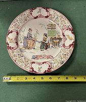 Front of the plate showing the decorative border and central illustration of two women with domestic items, along with the text 'A La Pantoufle de Cendrillon'. A measuring tape indicates approx 9 inches diameter.