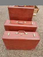 Three red-brown Samsonite suitcases arranged on gravel, front piece visible with gold-tone hardware and top handle