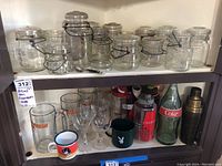 Shelf of glass jars with metal clasp lids (Atlas Ball canisters) on upper shelf. Lower shelf holds glassware including Hooters mugs, Playboy mug, Coca-Cola bottle, water bottles, and a metal shaker.