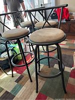 Three bar stools shown with black metal frame and beige ultra suede padded seats on a multicolored rug.
