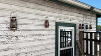 Six outdoor vintage-style metal lanterns hanging on a weathered white wooden wall, showing various degrees of rust and color variation.