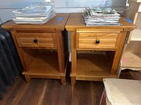 Two wooden nightstands side by side, both with one drawer and an open shelf below. Surfaces light to medium wood finish with visible grain.