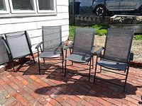 Four outdoor folding chairs arranged side by side on a brick patio, showing seating surface and armrests