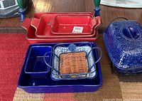 Assortment of red and blue ceramic roasting pans and baking dishes on a table including a ceramic dish with woven section.