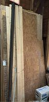Photo showing stacked plywood sheets and 2x4 lumber pieces leaning against a wall inside a shed.