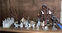 Wide view of assorted spooky plastic figurines on wooden shelf including gargoyle stone bridge, skeletons, witch, clown, headless horseman and trees.