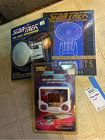 Photo showing the die-cast U.S.S. Enterprise starship model box, the Star Trek The Next Generation Enterprise blueprint booklet, and the Tiger Electronics Star Trek LCD video game in packaging.
