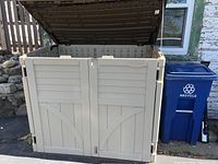 Front view of beige plastic Suncast storage box with lid open and blue recycle bin beside it