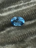 Close-up photo of the oval cut blue topaz gemstone on a textured fabric background showing facets and clarity.