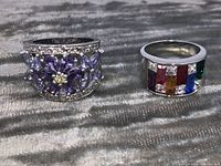 Two rings on gray textured fabric: left is purple stone floral design ring size 8.5, right is multi-colored stone ring size 7.
