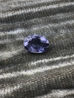 Close-up photo of blue oval faceted gemstone on textured fabric background showing multiple facets and blue color.