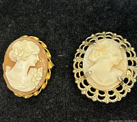 Two vintage cameo brooches on black background showing detailed carved female profiles and gold-tone frames.