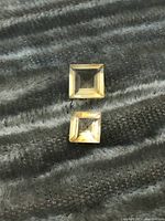 Two square-cut citrine gemstones placed side by side showing top view against a dark grey textured fabric background.