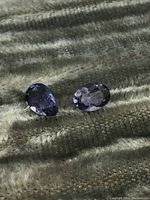 Two loose faceted Iolite gemstones placed on a textured dark fabric background, showing violet-blue color and oval and pear cuts.