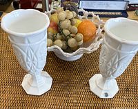 Two white milk glass chalice vases with geometric detail and octagonal bases shown on woven surface.