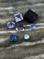 Six loose mixed cut gemstones on textured fabric background; various colors and cuts including square, oval, triangular, round.