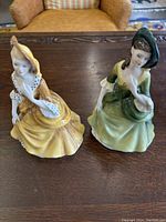 Two Royal Doulton porcelain figurines placed on a wooden table in front of a tan chair, showing full view of both figurines side by side.
