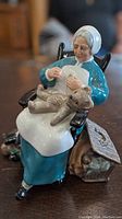 Front angled view of the Royal Doulton Nanny figurine seated in rocking chair, sewing a teddy bear, showing detailed facial expression and clothing.