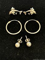 Three pairs of sterling silver earrings displayed on black fabric: dolphin-shaped studs, plain hoops, and small round studs.