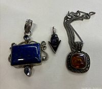 Photo of all three pendants: lapis lazuli rectangular pendant with silver accents, small amethyst pendant with V-shaped silver design, amber pendant with marcasite and chain.
