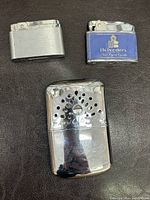 Photo showing two vintage lighters and one vintage hand warmer. All items appear metal with polished chrome-like finish. The hand warmer has decorative ventilation holes.