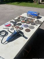 Overall view of the angle grinder and various discs spread out on a white folding table outdoors.