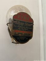 Photo showing clear glass fire grenade with metal label 'For Fire Shur-Stop The Automatic Fireman on the Wall' mounted in brass bracket.