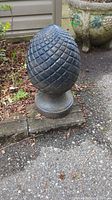 Antique French cast iron pinecone finial outdoors on a stone base near a planter