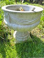 Side angle view showing the decorative garland design and weathered texture of the cement planter on a pedestal base outdoors