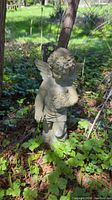 Full view of the concrete cherub statue standing outdoors surrounded by foliage, showing entire figure and wings.