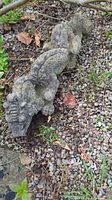 Full view of the concrete griffin gargoyle laying on gravel and garden soil showing length, texture, and form