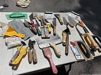 Overview of the full collection of drywall and tiling hand tools laid out on a table, showing various putty knives, scrapers, trowels, plastic spreader, and roller.