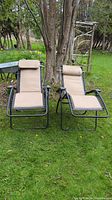 Two Lafuma gravity chairs with black metal frames and tan fabric seating positioned upright outdoors on grass near trees.