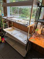 Front angle of white metal three-shelf hydroponic plant stand with trays showing dirt and residue, positioned near a window with natural light. Visible electrical cords and wiring for grow lights attached at the top shelf.