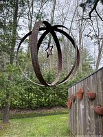 Photo of circular hanging metal garden art pendant suspended outdoors from a branch, showing five metal bands forming a spherical shape with a dangling star in the center. Background includes trees and garden area.