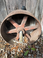 Iron garden art metal wheel resting against a wood fence with visible rust and faded red paint on six spokes and grooved rim.