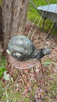 Close up front-side view of black metal snail on rusty trellis stand outdoors with wood and leaves background
