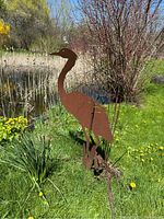 Side view of rust-colored metal crane garden art sculpture among grass and pond plants with bull rushes.