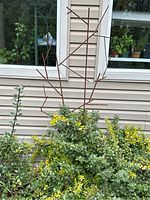 Two metal stick trellis sculptures placed in garden beds against a house siding. Rust-colored rods arranged to mimic branch structures.