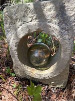 Stone piece with circular opening holding amber blown glass float, placed on garden ground near plants