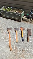 Four axes and one cleaver arranged on a gravel surface outdoors with planter and garden ornaments visible in the background.