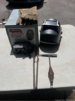 Photo showing Lincoln Electric Deluxe Flip Front Welding Helmet, gloves, goggles, tongs, and welding pick on a white table outdoors.