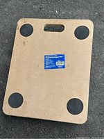 Top view of moving dolly showing rectangular wooden platform with label on top center and four black circular rubber pads on bottom corners.