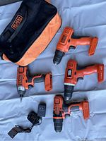Photo shows four cordless Black & Decker drills, a battery charger, and a branded soft carrying bag on a blue fabric surface.
